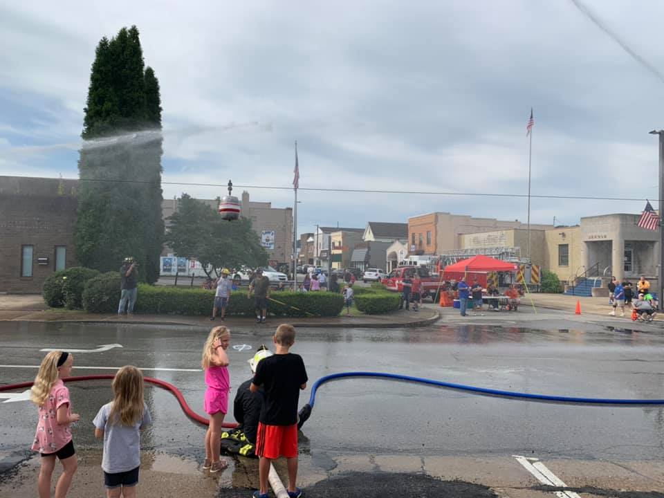 Children stand on a wet street holding hoses, spraying water as a firefighter demonstration takes place nearby. Fire trucks and onlookers are visible in the background, with buildings lining the small town street.